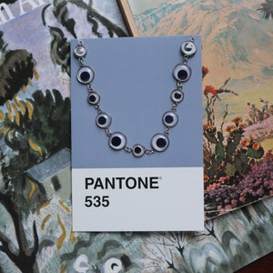 Può includere: Una collana color argento con pendenti circolari a forma di occhio, esposta su una scheda colore Pantone grigia con l'etichetta "535". La collana è ambientata su uno sfondo di libri d'arte vintage e una superficie di legno scuro.