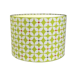 May include: A cylindrical lampshade featuring a vibrant green backdrop adorned with a repeating pattern of white floral designs. Each flower's center is accented with a small dot of colour, including red, blue, yellow, and grey. The lampshade is designed for a lamp base.