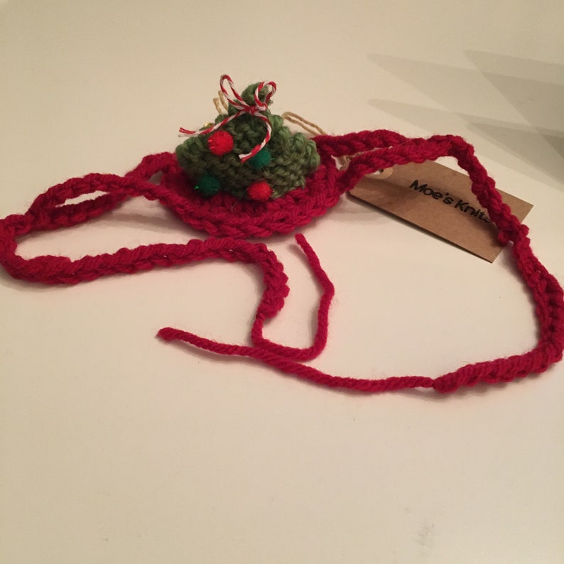 May include: A red crocheted headband featuring a small green crocheted Christmas tree ornament decorated with red and green pom-poms and a red and white twine bow. A brown cardboard tag with the text Moe's Knits is attached to the red band.