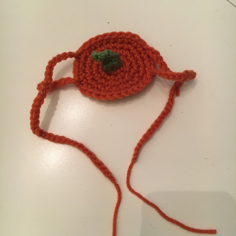 May include: A round, orange crocheted pumpkin accessory featuring a small, textured green stem at the center. The pumpkin is attached to two long, thin orange crocheted ties extending from the sides, designed for securing the item in place.