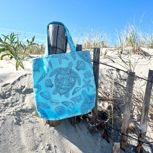May include: A light blue tote bag with a black and white design of sea turtles, seashells, and starfish. The bag is hanging on a wooden fence post against a backdrop of sand dunes and beach grass.