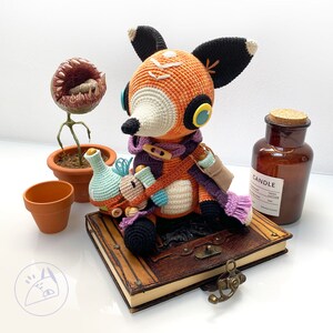 Peut inclure: Un renard en crochet orange avec une &eacute;charpe violette et un journal en cuir marron. Le renard tient une petite figurine en crochet. Le journal porte l'inscription "CANDLE FRESH EDITION" sur le c&ocirc;t&eacute;.