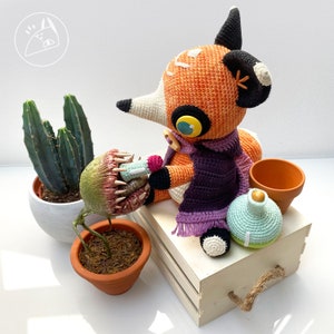 Peut inclure: Un renard en crochet portant une &eacute;charpe violette et tenant une petite plante en crochet. Le renard est assis sur une bo&icirc;te en bois avec une bouteille en crochet et une petite plante en pot.