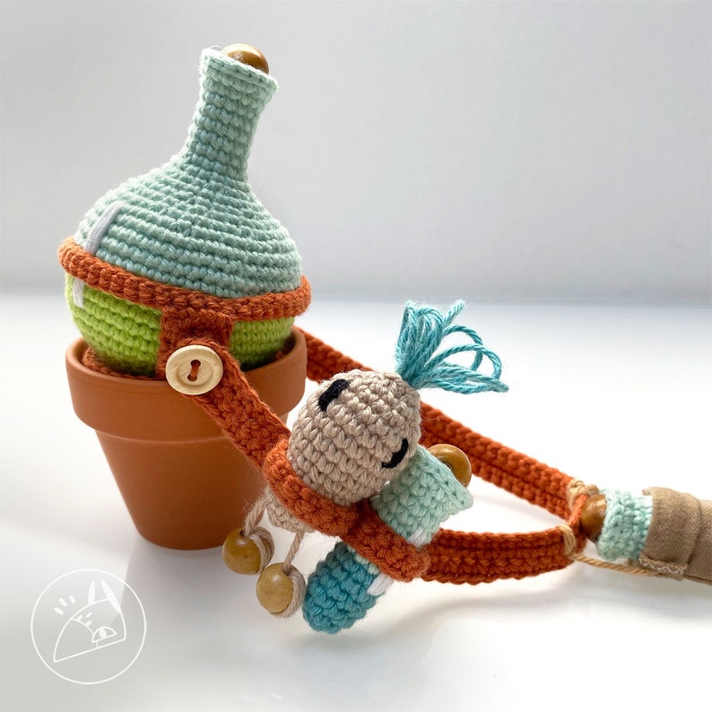 Peut inclure: Un jouet en crochet en forme de plante avec un pot vert, une tige brune et une fleur bleue. La plante est attach&eacute;e &agrave; une sangle brune avec une perle en bois.