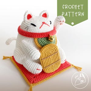 Puede incluir: Un gato Maneki Neko blanco de ganchillo con detalles rojos, un collar rojo y una campana dorada. El gato está sentado sobre una alfombra roja y amarilla. La imagen incluye el texto "CROCHET PATTERN".