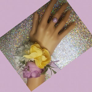 May include: A wrist corsage with yellow, white, and purple flowers. The corsage is tied with a white ribbon and is worn on a wrist with a purple ring.