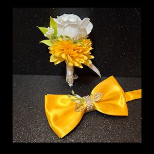 May include: A yellow bow tie with a rhinestone and leaf embellishment, and a boutonniere with a white rose and yellow chrysanthemums.