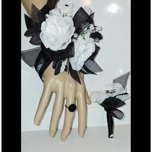 May include: A black and white floral wrist corsage and boutonniere set. The wrist corsage features white roses, black ribbon, and decorative accents. A matching black flower ring is also included. The boutonniere has white flowers and black accents.