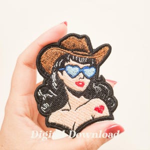 以下が含まれることがあります： 黒髪のカウガール、茶色のカウボーイハット、青いハート型のサングラスが特徴の刺繍パッチ。カウガールは赤いハートのタトゥーと赤い唇をしています。パッチは黒で縁取られています。
