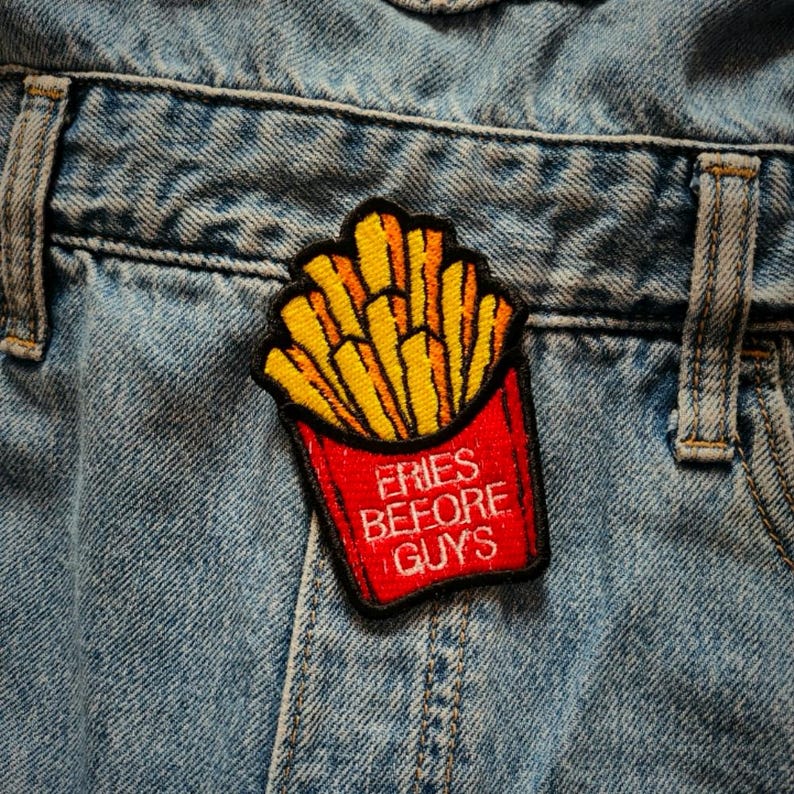 May include: A red and yellow embroidered patch with a cartoon image of french fries in a red container. The text "FRIES BEFORE GUYS" is written in white on the container.