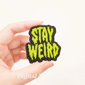 Op de afbeelding: Een geborduurde zwarte en groene patch met de tekst "Stay Weird" in een druppelvormig lettertype.