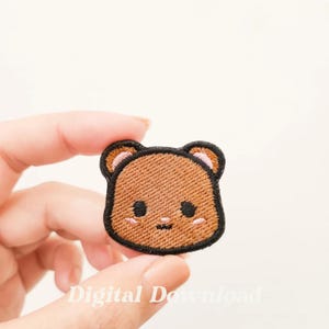 Könnte beinhalten: Ein braunes gesticktes Bärenkopf-Patch mit schwarzen Umrandungen und rosa Wangen. Das Patch wird von einer Hand gehalten.