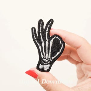 以下が含まれることがあります： スケルトンの手が「OK」サインをしている、黒い刺繍のアイロン接着パッチです。