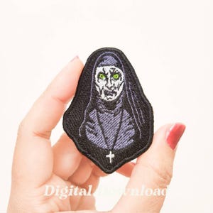 May include: Embroidered iron-on patch featuring a black and purple nun with green eyes. The nun has a cross on her chest.