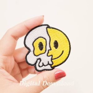 Pode incluir: Etiqueta bordada termocolante com uma caveira e um rosto sorridente, metade de cada desenho. A caveira é branca com contornos pretos e o rosto sorridente é amarelo com contornos pretos.