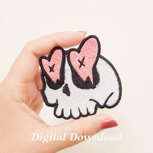 May include: White skull with pink hearts for eyes, each heart has a black X on it. The skull is outlined in black and has a black dripping effect from the eyes. The skull is embroidered on a white fabric.