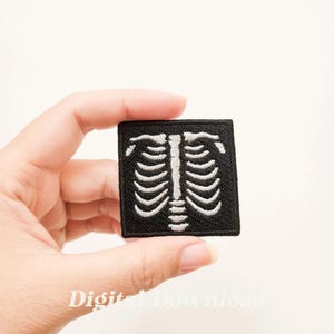 Puede incluir: Un parche bordado negro con un diseño de caja torácica de esqueleto blanco. El parche es de forma cuadrada y tiene un contorno blanco.