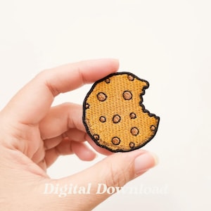 Pode incluir: Um remendo de biscoito bordado em marrom e amarelo com uma mordida. O remendo é contornado com fio preto.