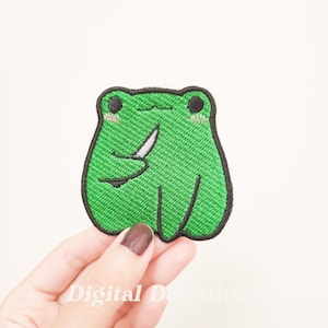 May include: Green embroidered iron-on patch of a frog holding a knife. The frog has a cute expression with big eyes and a small smile.