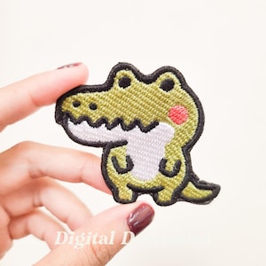 Può includere: Un toppa ricamata verde di un coccodrilo dei cartoni animati con la pancia bianca e un rossore rosso sulla guancia.