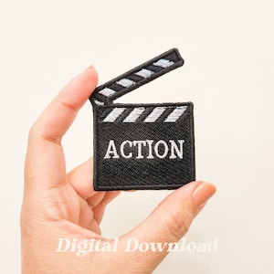 Puede incluir: Un parche de hierro bordado en blanco y negro de una claqueta de cine con la palabra "ACCIÓN" en texto blanco.