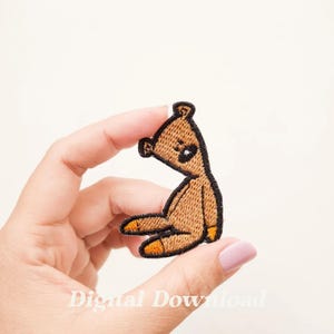 Könnte beinhalten: Ein gestickter Teddybär-Patch in Braun mit schwarzen Nähten und orangenen Füßen. Der Patch wird von einer Hand gehalten.