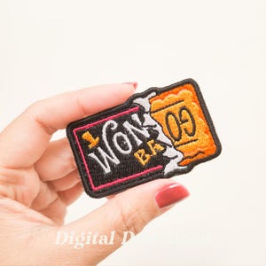 May include: Embroidered patch with the words "Won Bar" and "09" in a rectangular design. The patch features black, orange, white, and pink colors. The patch is held by a hand.