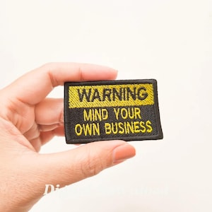 Könnte beinhalten: Ein gestickter schwarzer und gelber Aufnäher mit dem Text "WARNING MIND YOUR OWN BUSINESS".
