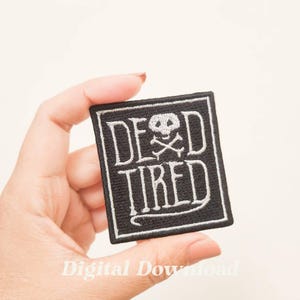 Op de afbeelding: Een zwarte vierkante patch met witte geborduurde tekst "DEAD TIRED". Een schedel en gekruiste botten grafiek staat boven het woord "TIRED". De patch heeft een witte rand.