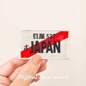 May include: Rectangular embroidered patch featuring the word "JAPAN" in black, a red diagonal stripe, and Japanese characters. The patch has a cream background and a stitched border.