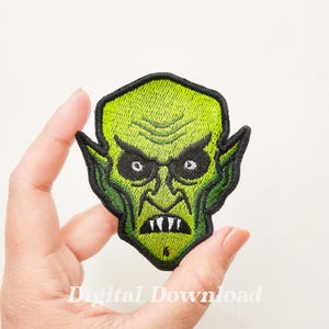 May include: Green and black embroidered patch of a monster's face with sharp teeth and large eyes.