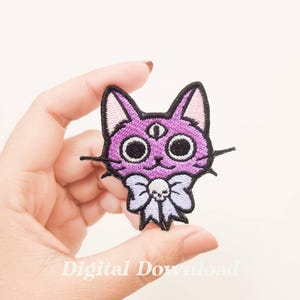 May include: Purple embroidered iron-on patch of a cat with a third eye and a bow tie. The bow tie has a skull and crossbones design.