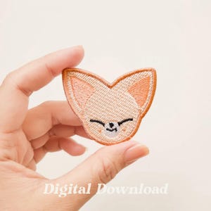 Puede incluir: Un parche bordado con la cara de un zorro con los ojos cerrados y una pequeña sonrisa. El zorro tiene orejas de color melocotón y una nariz blanca.