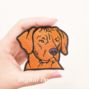 Op de afbeelding: Geborduurde patch met een hondenhoofd in oranje en zwart. De hond heeft een gedetailleerd ontwerp met een zwarte neus en ogen. De patch is waarschijnlijk voor kleding of accessoires.