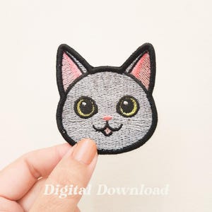 Könnte beinhalten: Gestickter Bügel-Patch mit einem grauen Katzenkopf mit schwarzen Ohren, rosa Wangen und gelben Augen. Die Katze hat eine kleine rosa Nase und einen lächelnden Mund.