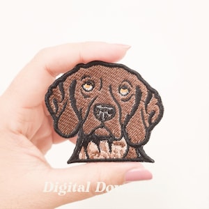 Op de afbeelding: Geborduurde patch met een bruine hond met een zwarte omtrek. De hond heeft gele ogen en een zwarte neus. De patch wordt in een hand gehouden, met de tekst "Digital Dow" onderaan. Decoratief item.