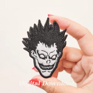 May include: Black and white embroidered patch featuring a character with spiky black hair and a wide, toothy grin. The character's eyes are wide open and have a menacing look.