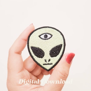 Op de afbeelding: Groene alien-gezichts-gebreide patch met een derde oog in het midden van het voorhoofd. De patch heeft zwarte stiksels en een zwarte omtrek.