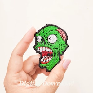 Puede incluir: Parche bordado de cabeza de zombie verde con un cerebro rosa, ojos blancos y una boca abierta con dientes afilados.