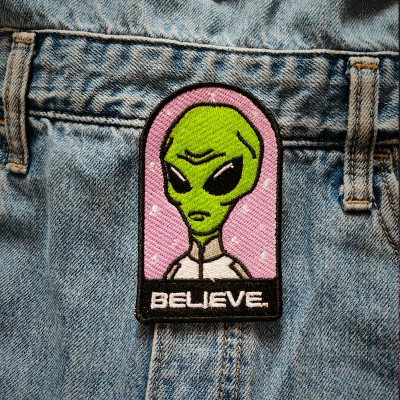 May include: A green alien with large eyes and a serious expression is featured on a pink and white embroidered patch. The patch has a black border with the word "BELIEVE" in white lettering.