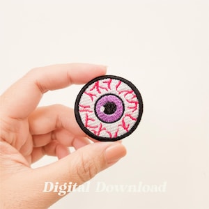 Op de afbeelding: Een ronde, witte, geborduurde patch met een roze oog en rode aderen. Het oog heeft een zwarte pupil en een paarse iris.