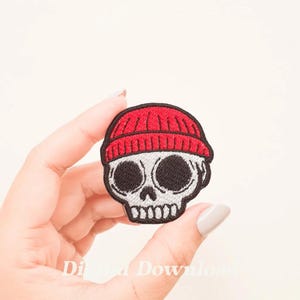May include: A white embroidered skull wearing a red beanie. The skull has black eyes and a small mouth.