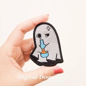 Op de afbeelding: Geborduurde patch met een cartoongeest die uit een blauwe fles drinkt. De geest is grijs met zwarte accenten en een kruis op zijn hoofd. De patch wordt door een hand vastgehouden.
