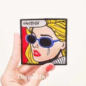 May include: Embroidered patch featuring a pop art style illustration of a blonde woman wearing blue sunglasses and crying. A speech bubble above the woman's head says "WHATEVER". The patch has a red and black border.