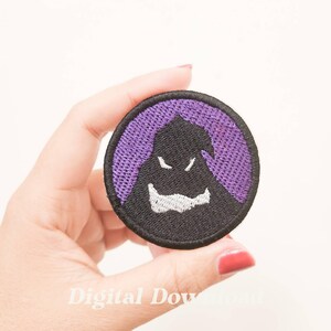 May include: A purple and black embroidered iron-on patch featuring a cartoon ghost with a mischievous grin.
