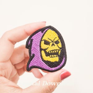 May include: Purple and yellow embroidered iron-on patch featuring a cartoon skull with a purple hooded robe.