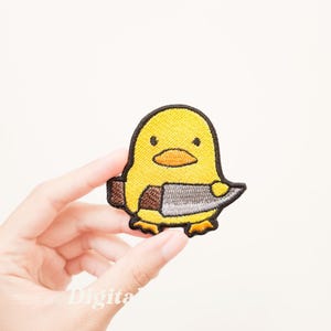 May include: Embroidered patch featuring a yellow cartoon chick holding a silver knife. The chick has a black outline, orange beak, and small black eyes. The knife has a brown handle. The patch is held in a hand against a white background.