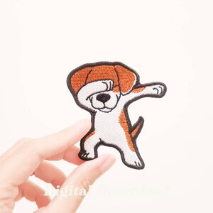 May include: Embroidered iron-on patch of a brown and white dog doing the dab dance. The dog has a brown head and a white body with brown spots. The patch is on a white background.