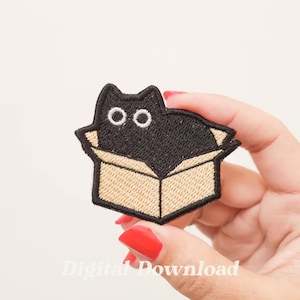 Op de afbeelding: Gestikte patch van een zwarte kat met witte ogen, zittend in een bruine kartonnen doos. De patch wordt vastgehouden in een hand met rode nagellak.