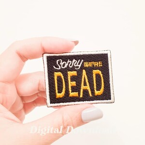 Puede incluir: Un parche bordado negro con costuras blancas alrededor de los bordes. El parche dice "Lo siento, estamos muertos" en letras amarillas.